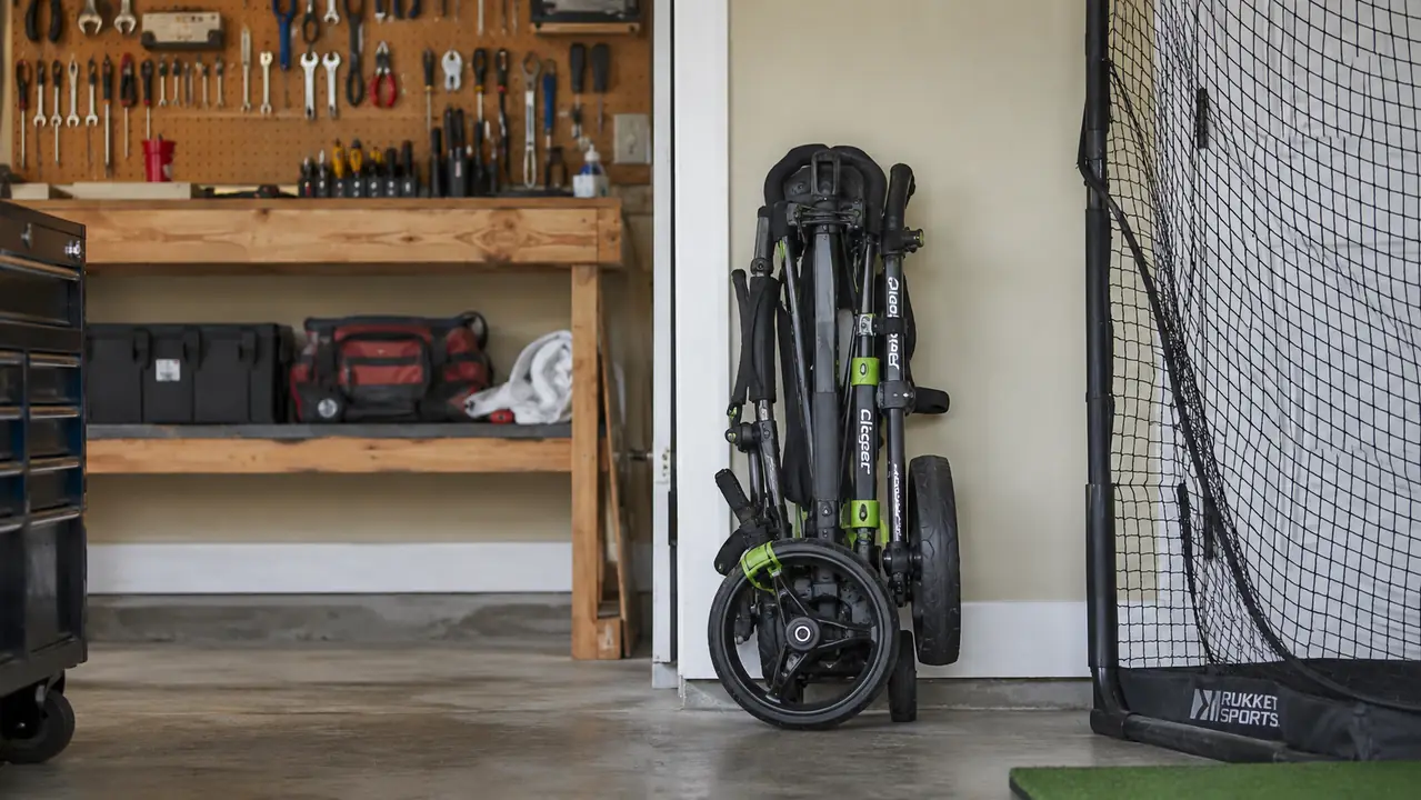 A three-wheel golf push cart folded flat and standing upright against a garage wall next to a practice net